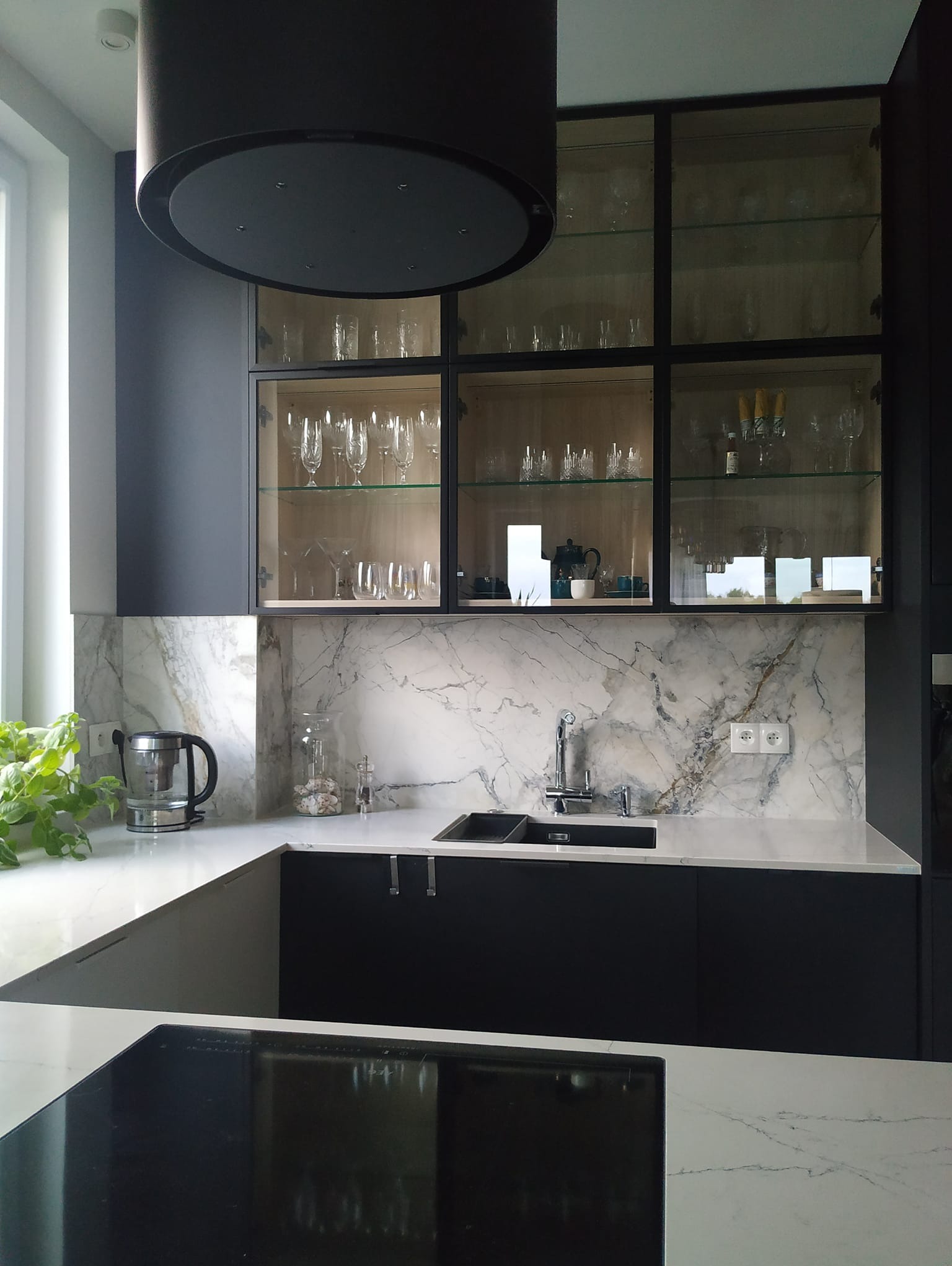 Nowoczesna kuchnia z czarnymi szafkami i blatem z białego marmuru. Nad blatem szafki z przeszklonymi drzwiami, w których widoczne są szklanki i naczynia.