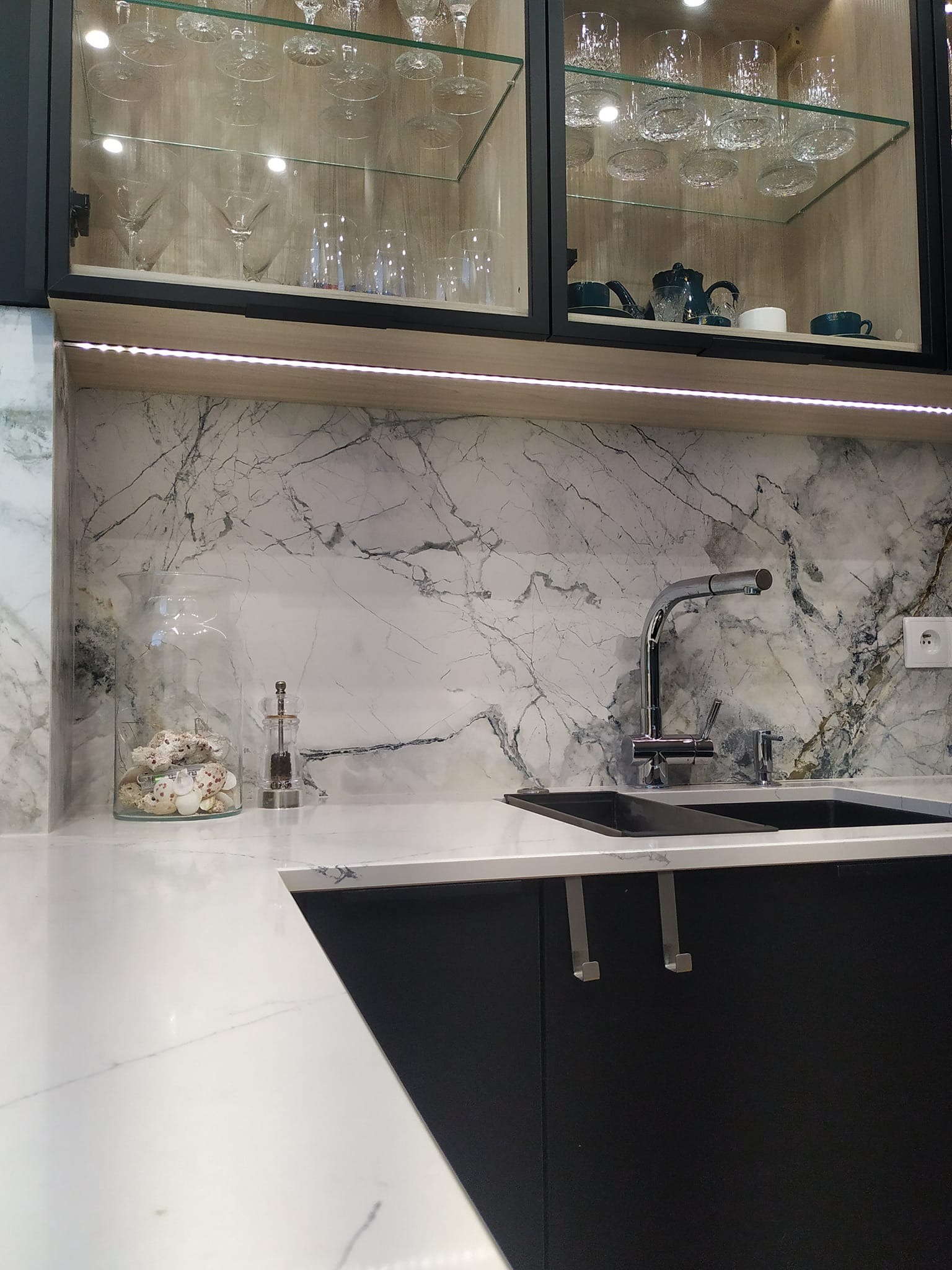 Nowoczesna kuchnia z marmurowym blatem i szafkami z ekspozycją szkła. Bateria kuchenna i zlewozmywak wkomponowane w elegancki design. Dekoracje z muszelek w szklanym naczyniu.
