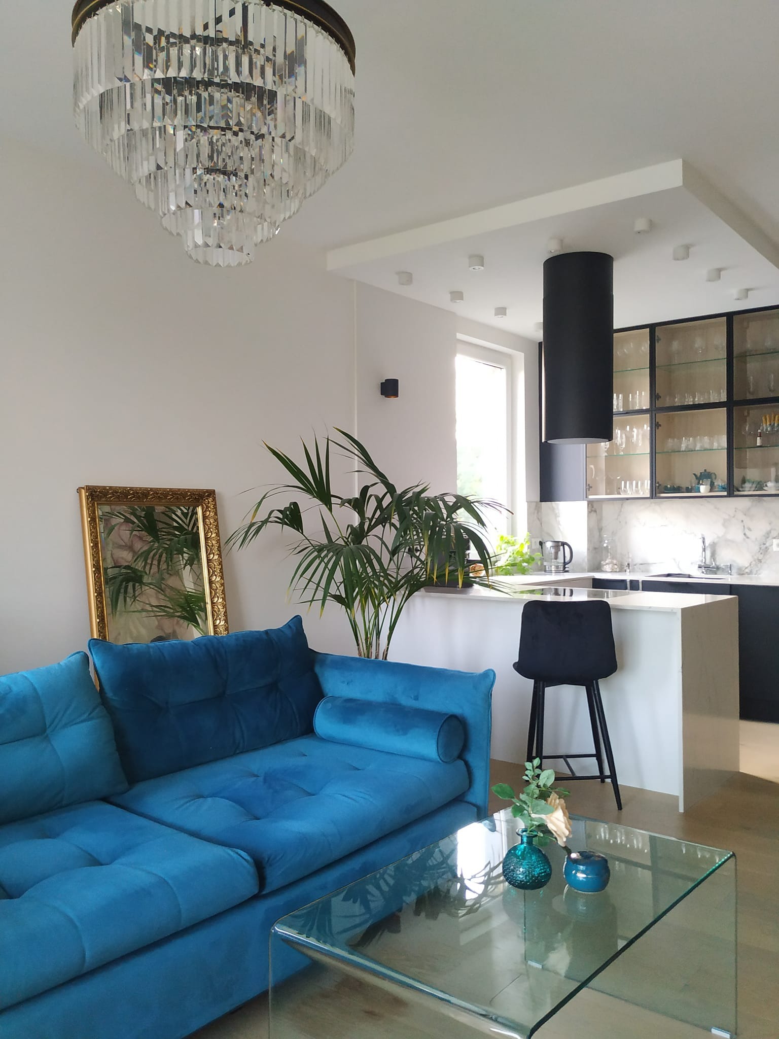 Salon z turkusową sofą, szklanym stolikiem i roślinami. W tle kuchnia z czarnymi szafkami i marmurowym blatem. Nad sofą kryształowy żyrandol.