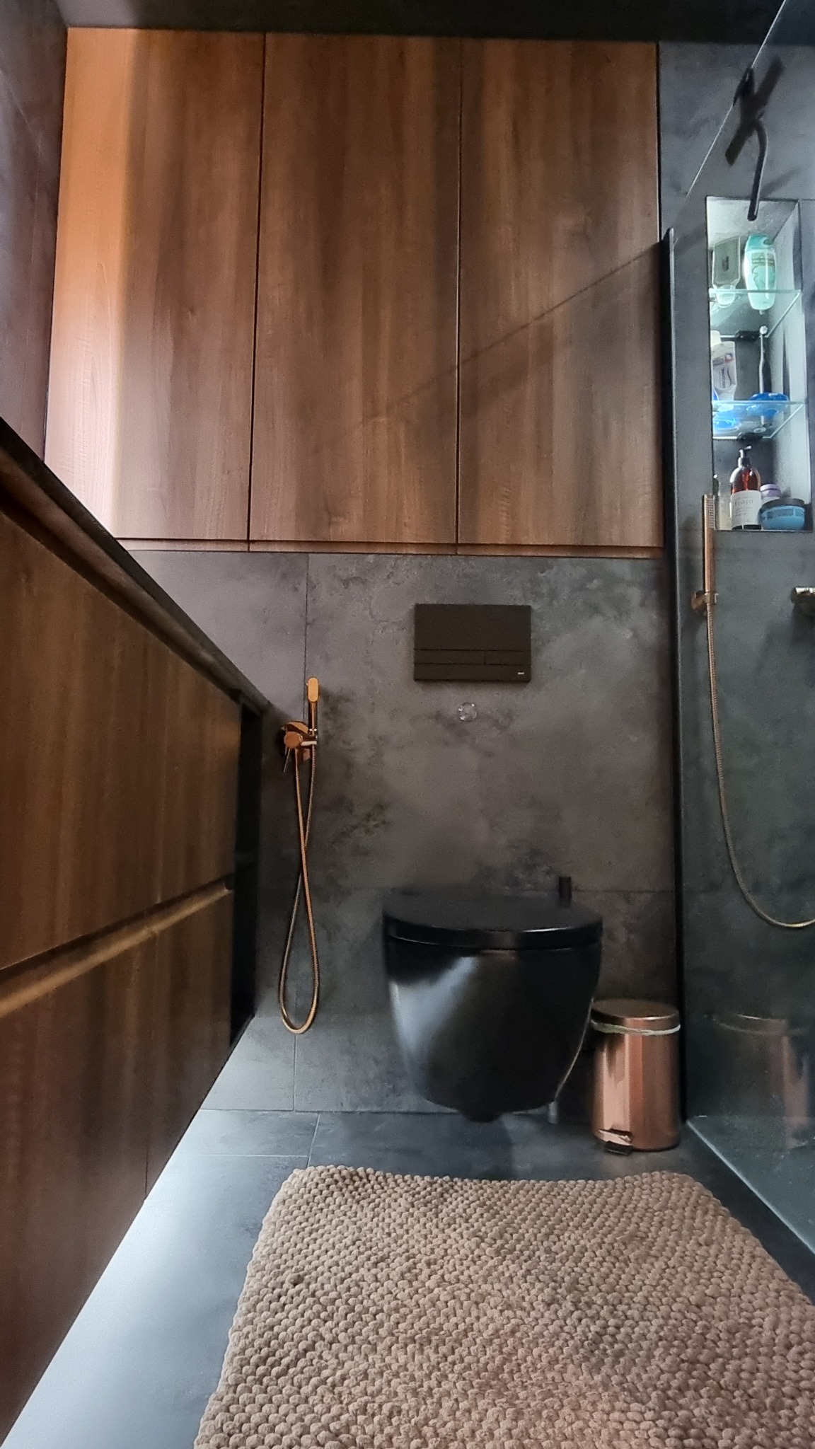 Nowoczesna łazienka z wiszącą toaletą w kolorze czarnym, armaturą w odcieniach miedzi i drewnianymi szafkami. Na podłodze beżowy dywanik.