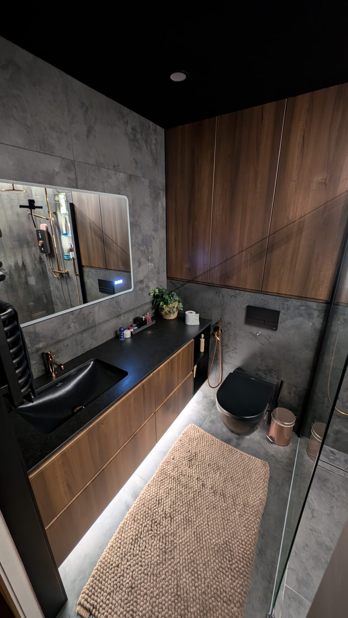 Nowoczesna łazienka z szarą płytką, drewnianymi szafkami pod umywalką z podświetleniem LED, czarną armaturą i dywanikiem. Minimalistyczny design.