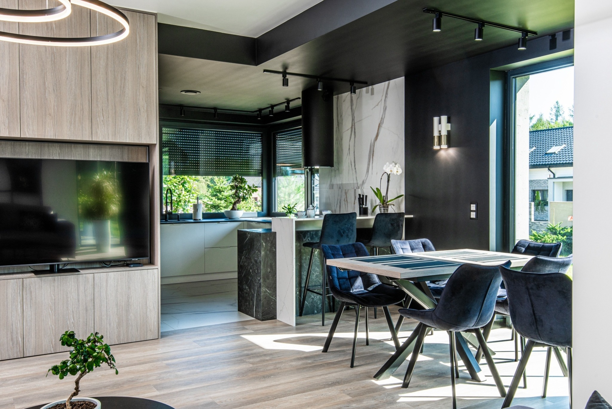 Nowoczesne wnętrze: jadalnia z ciemnymi krzesłami i drewnianym stołem, widok na kuchnię z marmurowym blatem i oknem na zieleń. Czarny sufit z oświetleniem punktowym.