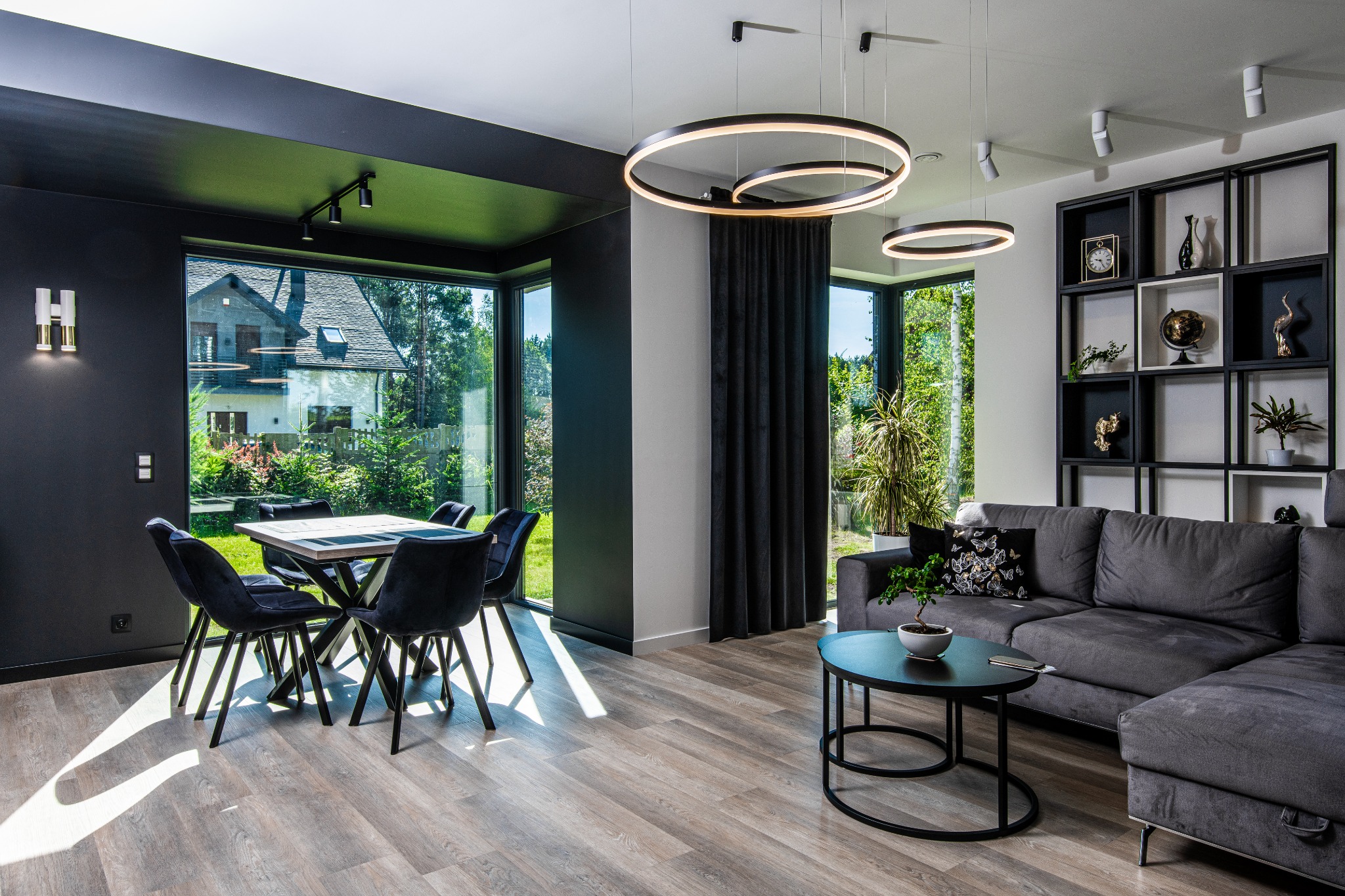 Nowoczesny salon z dużym oknem, czarnymi ścianami i okrągłymi lampami. W tle widoczny dom przez okno, a w pomieszczeniu stół z krzesłami i szara sofa.