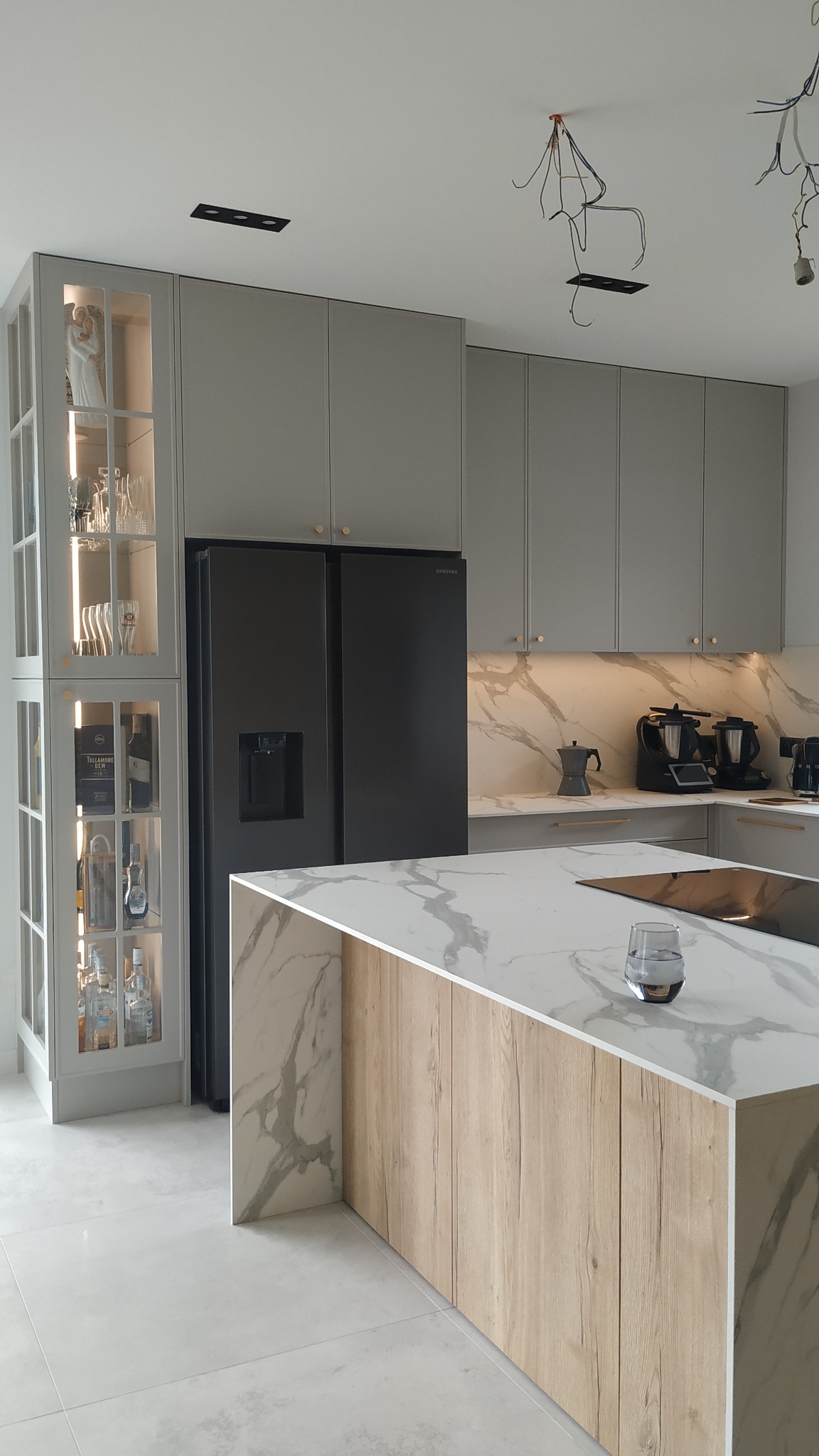 Nowoczesna kuchnia z wyspą, szare szafki, marmurowy blat i backsplash, czarna lodówka Samsung. Widoczne niedokończone oświetlenie sufitowe.