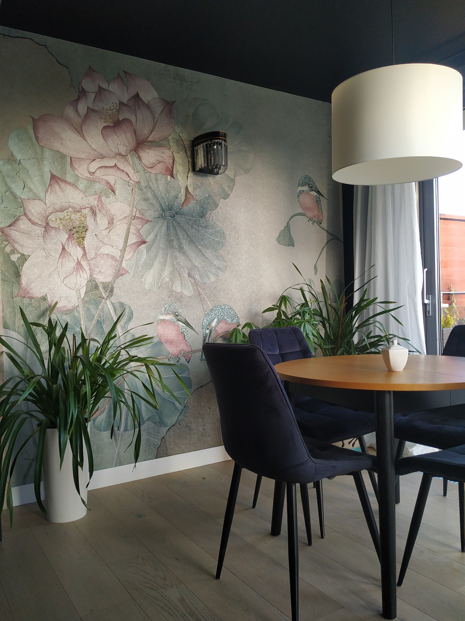 Jadalnia z okrągłym stołem i krzesłami na tle ściany z motywem kwiatowym i ptaków. Nowoczesna lampa wisząca, rośliny doniczkowe. Parkiet na podłodze.