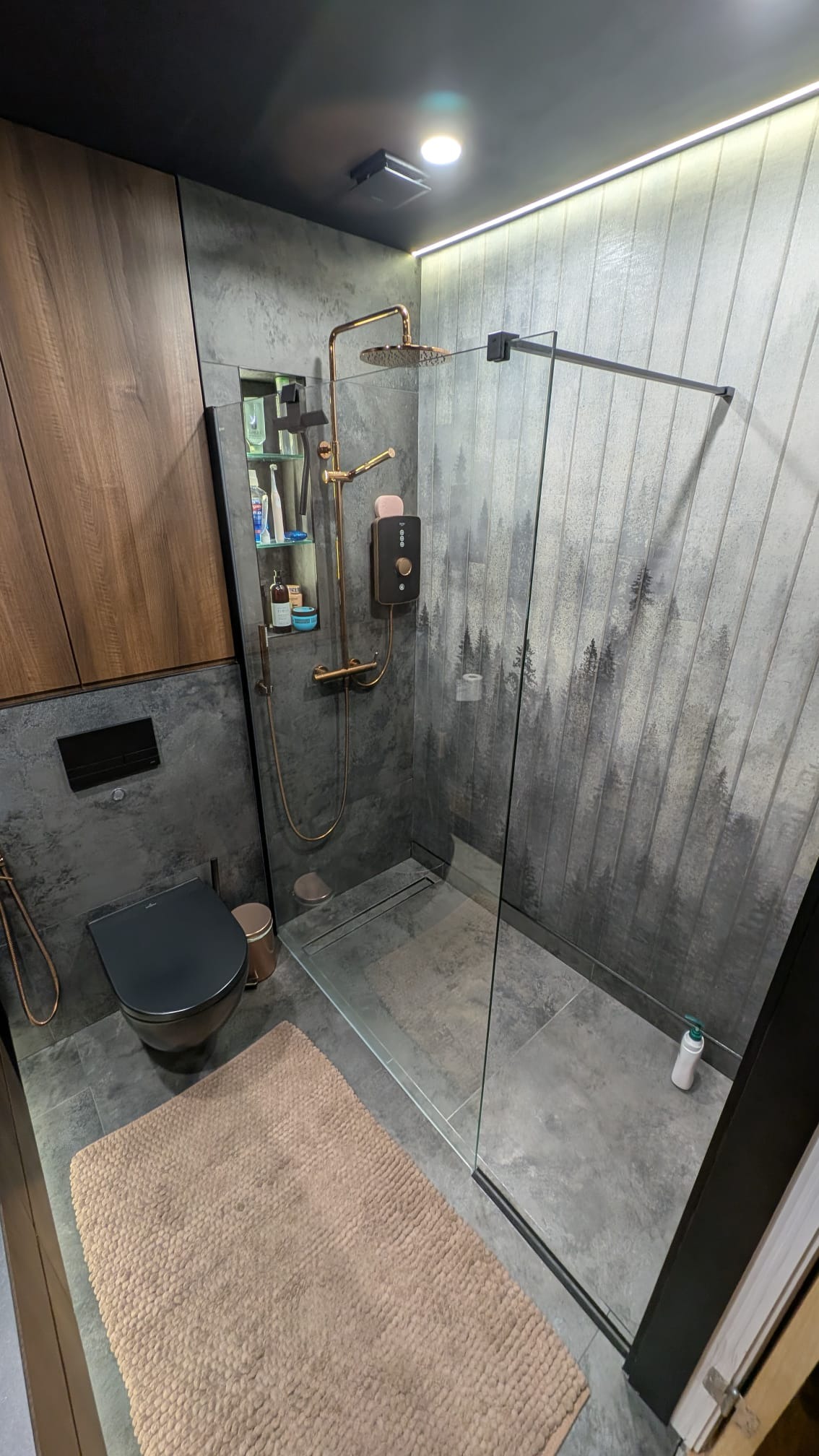 Nowoczesna łazienka z kabiną prysznicową typu walk-in, szarą armaturą i płytkami z motywem lasu. Uwagę przykuwa złota deszczownica i minimalistyczna toaleta.