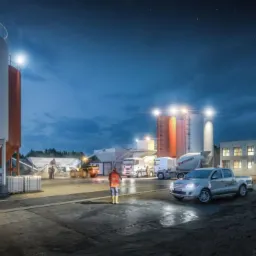Zakład produkcji betonu oświetlony nocą, z widocznymi silosami, betoniarkami, pickupem z logo AKJ i pracownikiem w kamizelce odblaskowej.