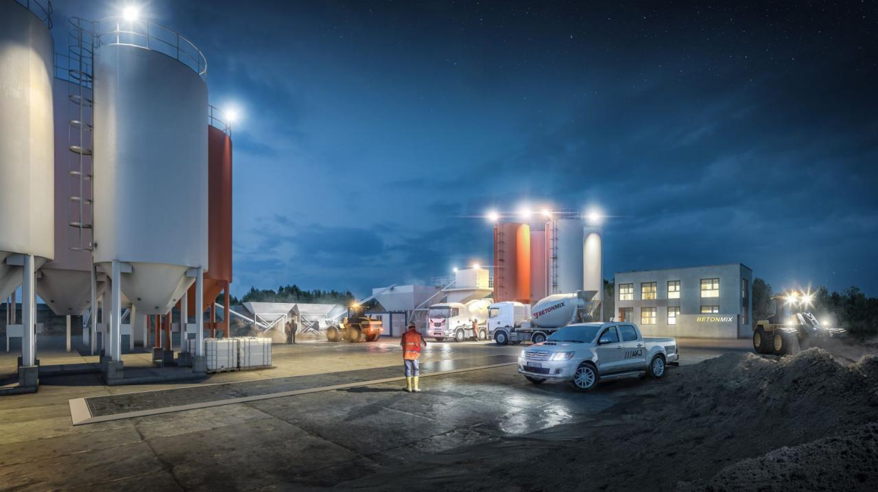 Zakład produkcji betonu oświetlony nocą, z widocznymi silosami, betoniarkami, pickupem z logo AKJ i pracownikiem w kamizelce odblaskowej.