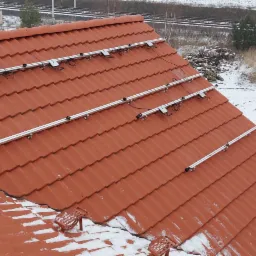 Montaż szyn do paneli fotowoltaicznych na dachu z czerwonej dachówki w zimowy dzień, widoczne uchwyty i kable, w tle ośnieżone pole, drzewa i linia kolejowa.