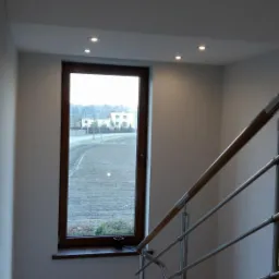 Wnętrze domu w Radostowicach po remoncie: widok na schody z drewnianą balustradą i metalowymi elementami, parkiet oraz okno z widokiem na zewnątrz.