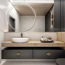 Nowoczesna łazienka z okrągłym lustrem podświetlonym LED, drewnianym blatem, białą umywalką nablatową i szufladami w kolorze grafitowym z okrągłymi, złotymi uchwytami. Na blacie zestaw kosmetyków...