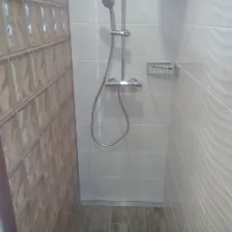 Wykończona kabina prysznicowa z szklanymi luksferami, białe płytki z falistą strukturą, szara podłoga imitująca drewno oraz srebrna bateria i półka na kosmetyki.