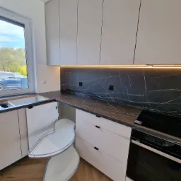 Nowoczesna kuchnia z białymi frontami, czarnym blatem imitującym marmur i wysuwaną półką narożną typu 'nerka', oświetlona listwą LED pod szafkami wiszącymi.