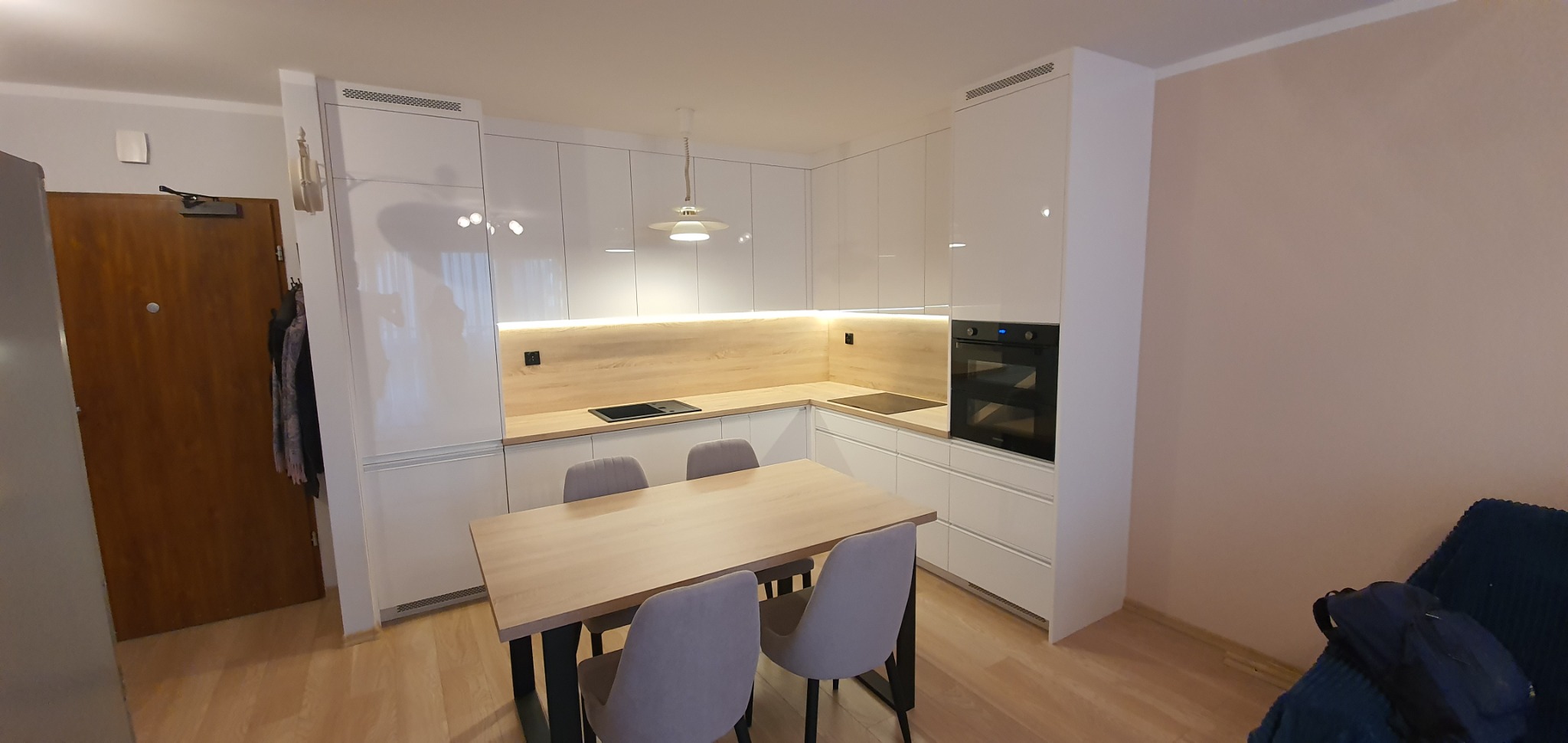 Jasna kuchnia z białymi, błyszczącymi frontami szafek, drewnianym blatem i podświetleniem LED, uzupełniona o stół z czterema szarymi krzesłami, widoczny w tle fragment pokoju z drewnianymi drzwiami.