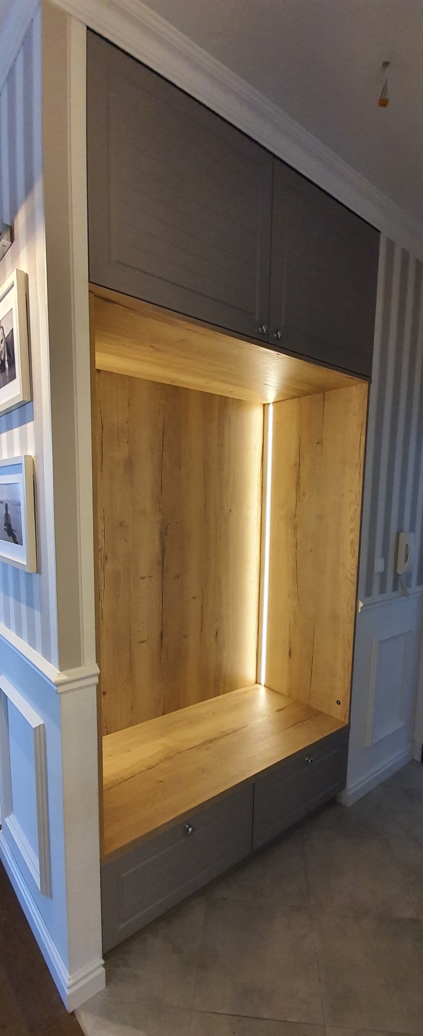 Wnęka z szarymi szafkami, drewnianym wnętrzem i oświetleniem LED, wkomponowana w ścianę z tapetą w paski.