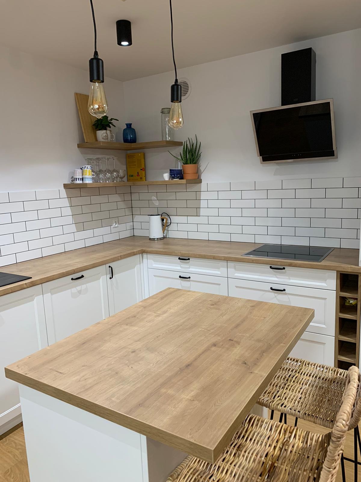 Biała kuchnia z drewnianym blatem i wyspą, oświetlona wiszącymi lampami z żarówkami Edisona, uzupełniona o drewniane półki z dekoracjami i białą cegłę na ścianie.