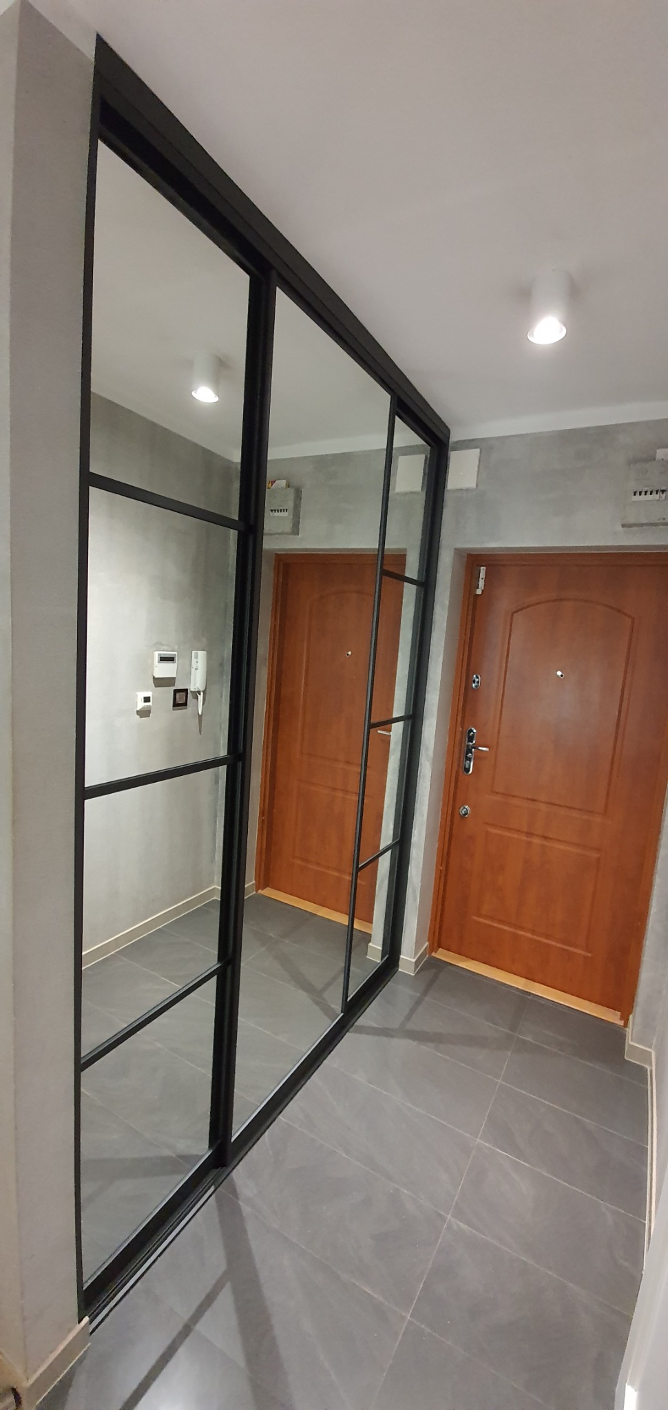 Szafa wnękowa z przesuwnymi drzwiami lustrzanymi w czarnej ramie, z podziałem na sekcje, w korytarzu z szarą podłogą i dwojgiem drewnianych drzwi wejściowych w tle.