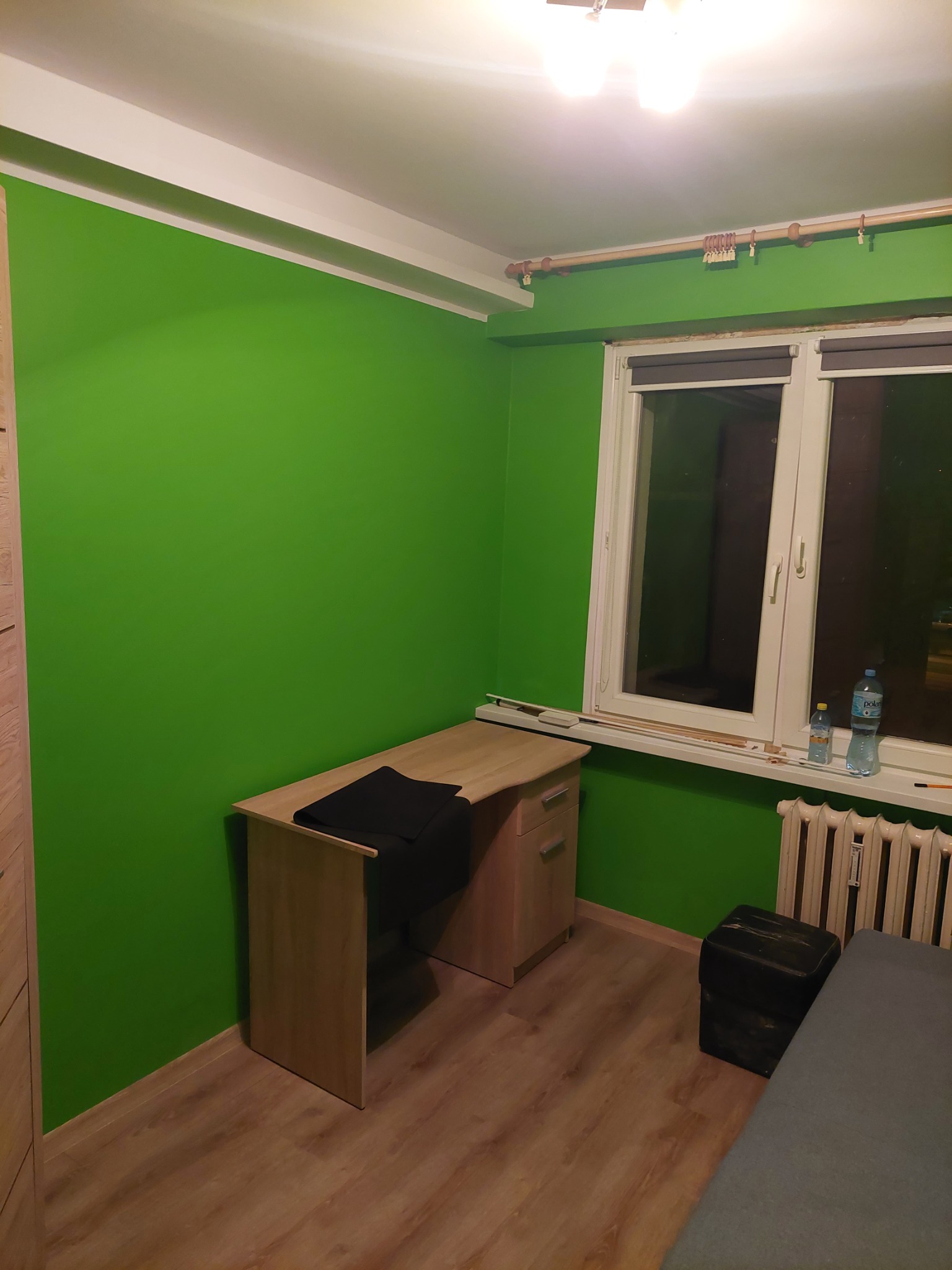 Świeżo pomalowany pokój z intensywnie zieloną ścianą, biurkiem i grzejnikiem pod oknem, widoczne ślady po remoncie na parapecie.