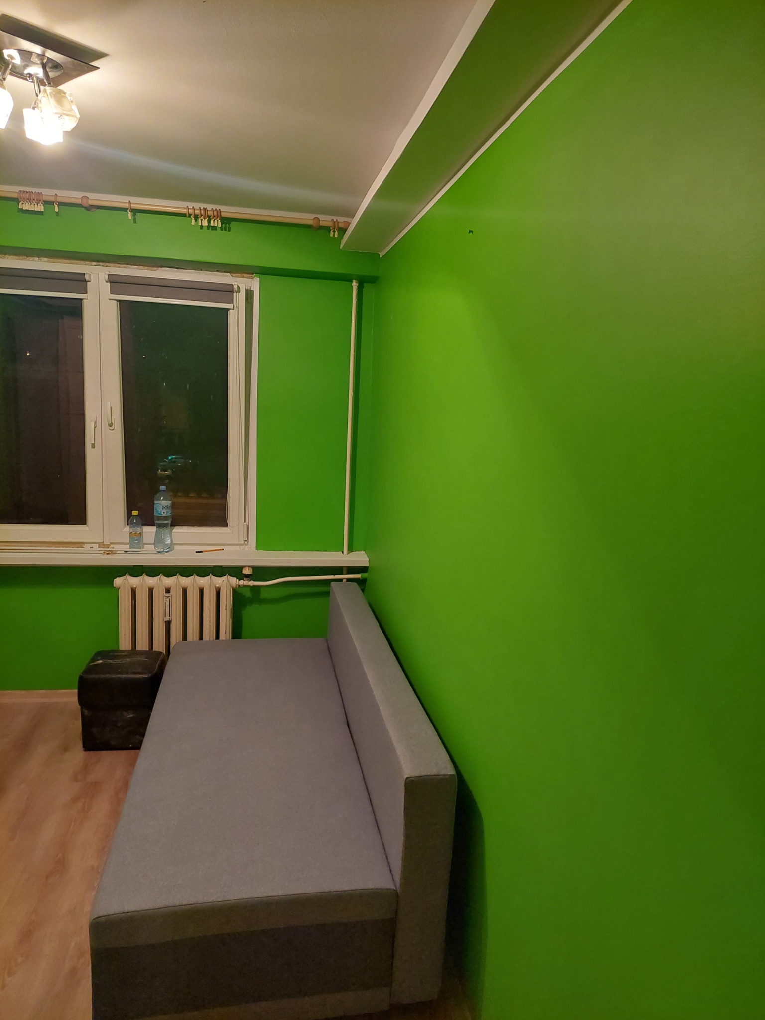 Ściana pomalowana na jaskrawy zielony kolor po wykonaniu prac w Białymstoku, widoczny fragment szarej sofy i okno z butelkami wody.