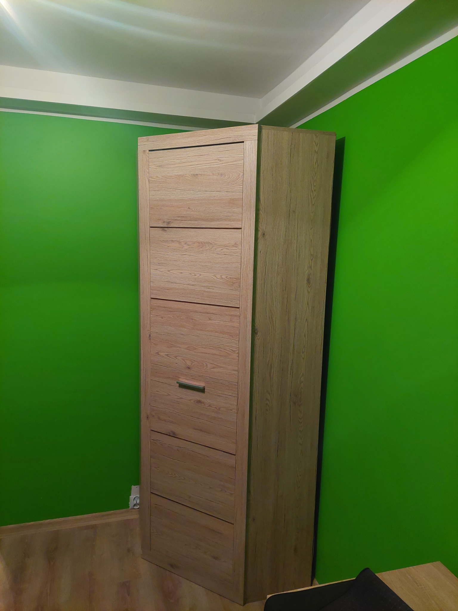 Wysoka, wąska szafka na buty z drewnopodobnym wykończeniem, ustawiona przy intensywnie zielonej ścianie. Widoczny kontakt elektryczny przy podłodze.