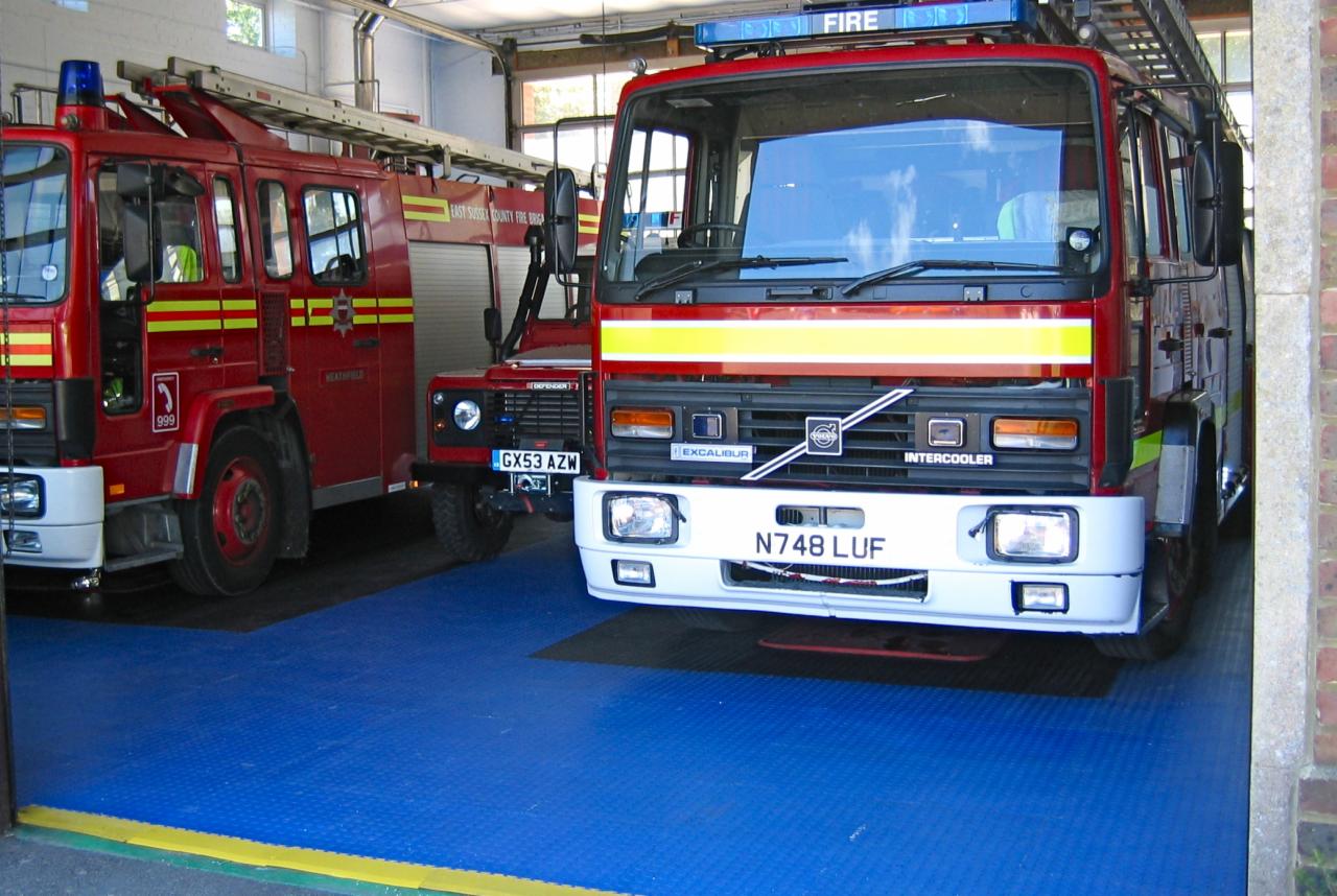 Dwa czerwone wozy strażackie zaparkowane w garażu na niebieskiej, modułowej podłodze z tworzywa sztucznego.