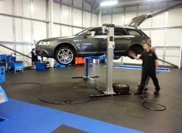 Szary samochód kombi na podnośniku w warsztacie. Mężczyzna w czarnym uniformie stoi obok, trzymając kabel. W tle widoczne narzędzia i wyposażenie warsztatowe, oraz podłoga w kratkę w kolorze czarnym.