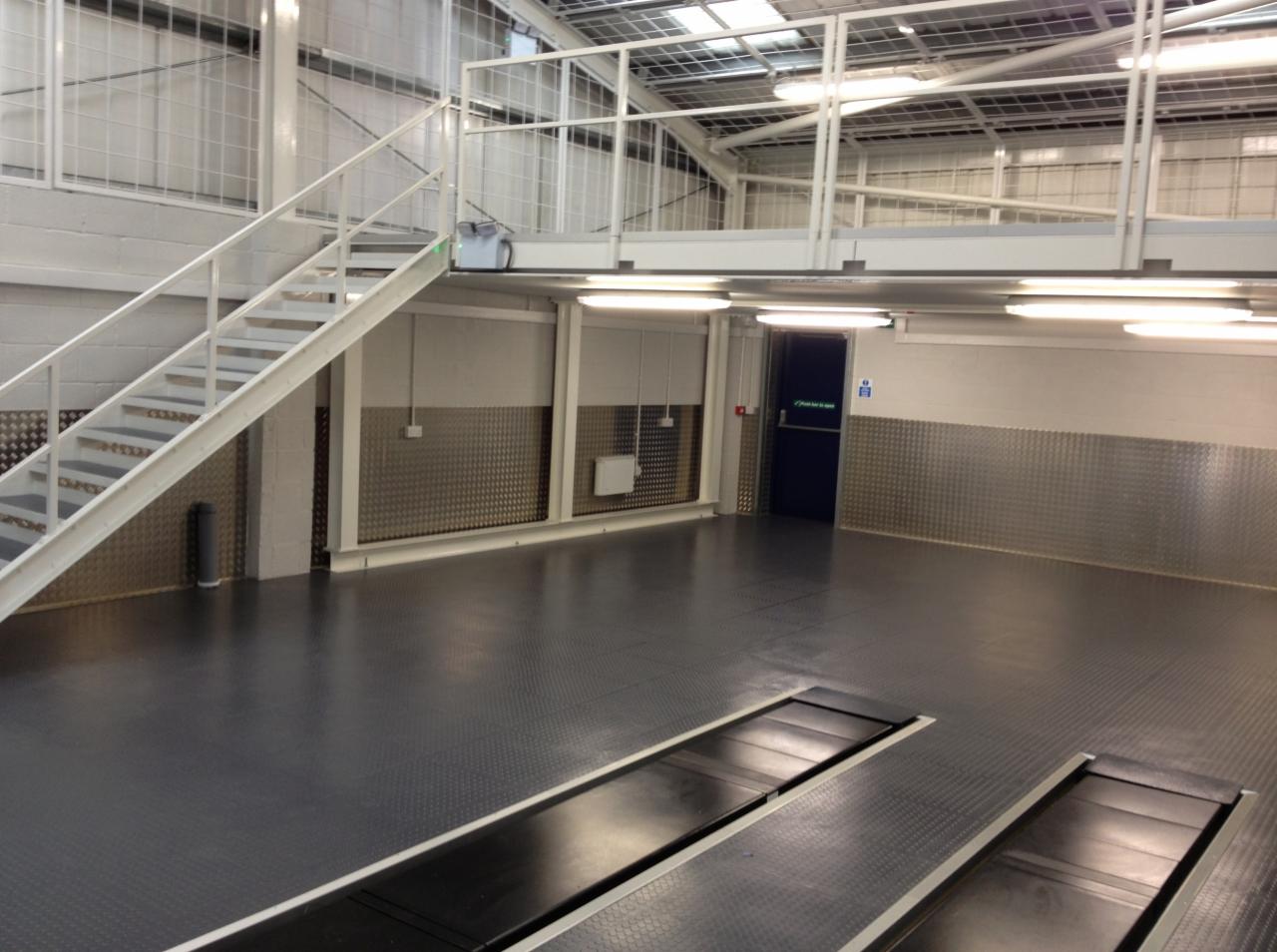 Wnętrze hali przemysłowej z szarą, antypoślizgową podłogą z fakturą, widoczne kanały rewizyjne, metalowe panele ścienne, schody i antresola, oświetlenie jarzeniowe.