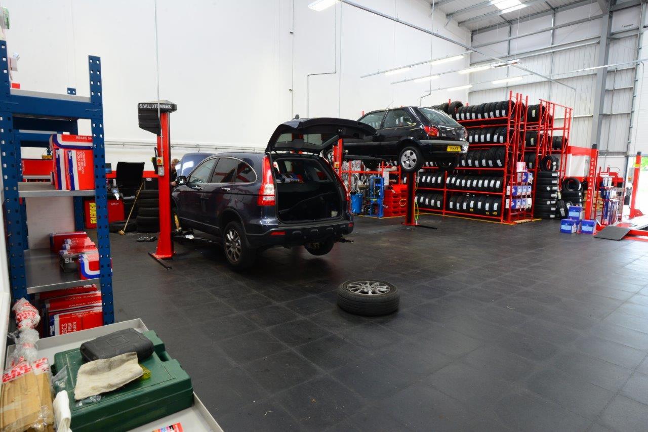 Wnętrze warsztatu samochodowego w Luton z dwoma samochodami na podnośnikach, regałami z oponami i czarną posadzką z gumy.