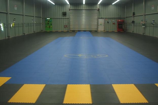 Wnętrze hali produkcyjnej z podłogą z modułowych płyt w kolorach niebieskim, szarym i żółtym. Na niebieskiej części widoczne logo. W tle zamknięta brama wjazdowa i metalowe ściany.