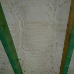 Poddasze z widoczną strukturą natryskowej izolacji termicznej, z fragmentami drewnianej konstrukcji więźby dachowej pomalowanej na zielono.