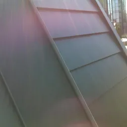 Fragment dachu pokrytego ciemnoszarą, metalową dachówką modułową, widoczny pod kątem z elementami konstrukcyjnymi, w tle budynki.