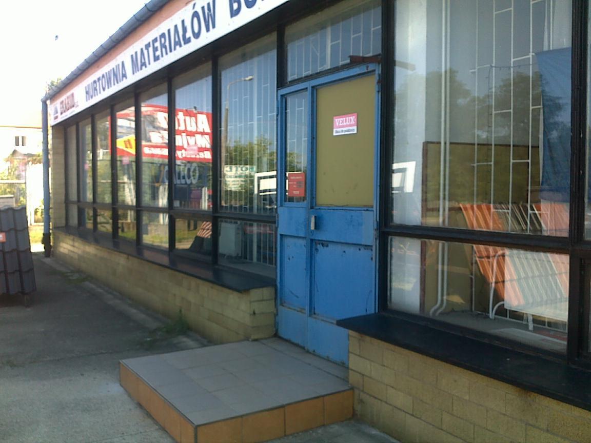 Fasada budynku hurtowni materiałów budowlanych z niebieskimi drzwiami wejściowymi i dużymi oknami wystawowymi, w których odbija się otoczenie.