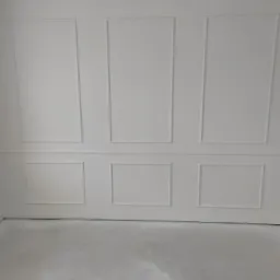 Biała ściana z dekoracyjnymi panelami w stylu angielskim, widoczne prostokątne ramy, szare podłoże, minimalizm.