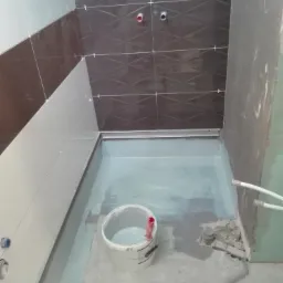 Wnętrze łazienki w trakcie remontu: ułożone płytki na ścianach z poziomym pasem brązowych płytek z wzorem, podłoga pokryta warstwą hydroizolacji, wiadro z farbą w rogu.