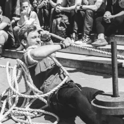 Czarno-białe zdjęcie mężczyzny ciągnącego linę przymocowaną do ciężaru, z publicznością obserwującą na schodach w tle.