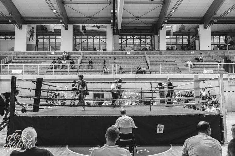 Czarno-białe zdjęcie ringu bokserskiego podczas zawodów, z widownią w tle i sędzią w ringu.