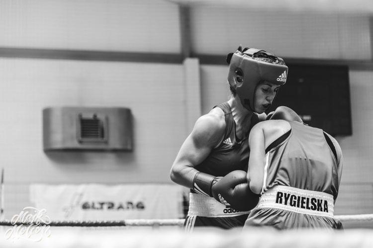 Czarno-białe zdjęcie z ringu bokserskiego: kobieta w kasku i rękawicach bokserskich w zwarciu z przeciwniczką, widoczny napis 'RYGIELSKA' na spodenkach, tło stanowią ściany hali sportowej.