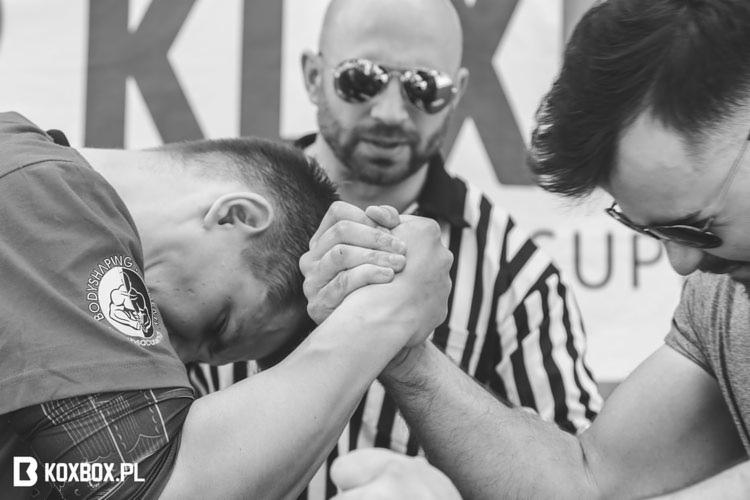 Czarno-białe zdjęcie dwóch mężczyzn rywalizujących w siłowaniu na rękę, z sędzią w tle, wszyscy w okularach przeciwsłonecznych.