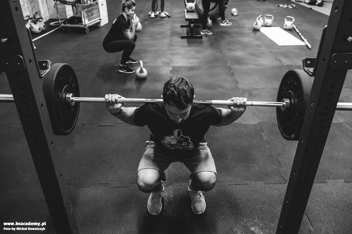 Czarno-białe zdjęcie mężczyzny wykonującego przysiad ze sztangą w siłowni, z widocznymi kettlebells i innymi osobami ćwiczącymi w tle.