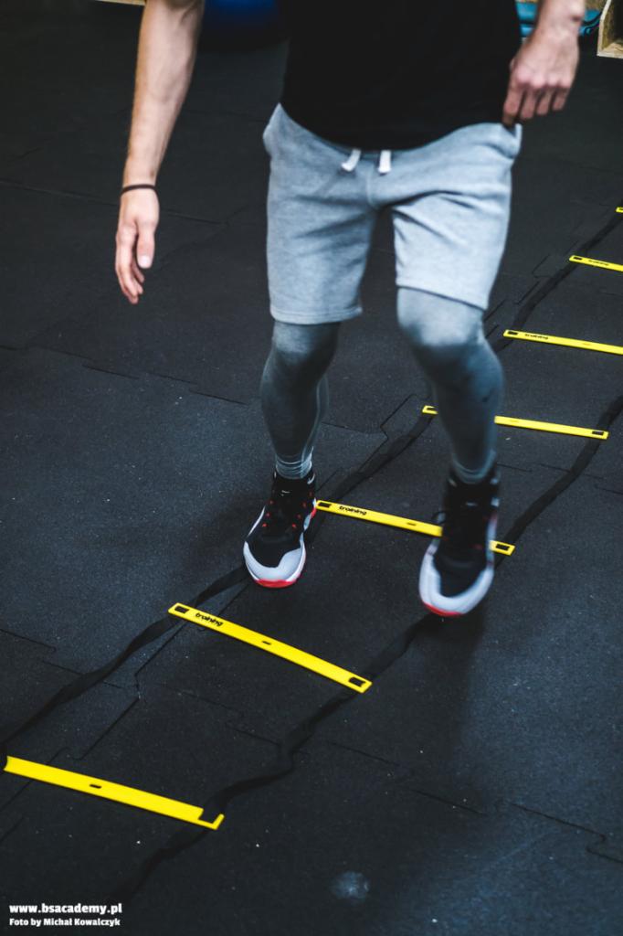 Osoba w szarych spodenkach i legginsach ćwiczy koordynację na czarnej podłodze z żółtą drabinką treningową; widoczne stopy w sportowych butach.