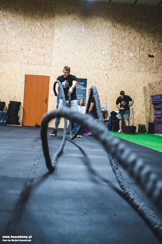Mężczyzna w czarnej koszulce ćwiczy z grubymi linami w siłowni, w tle widoczni inni ludzie i tablica z rozpiską ćwiczeń.