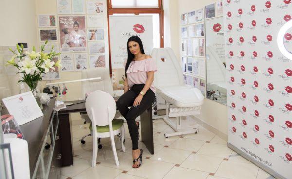 Kobieta w różowej bluzce i czarnych spodniach siedzi na krześle w jasnym salonie kosmetycznym z białym fotelem zabiegowym w tle, ściany zdobią plakaty i dyplomy.