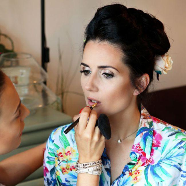 Kobieta w kwiecistej koszuli otrzymuje aplikację szminki pędzelkiem, widoczny delikatny makijaż oczu i upięcie włosów z kwiatem.
