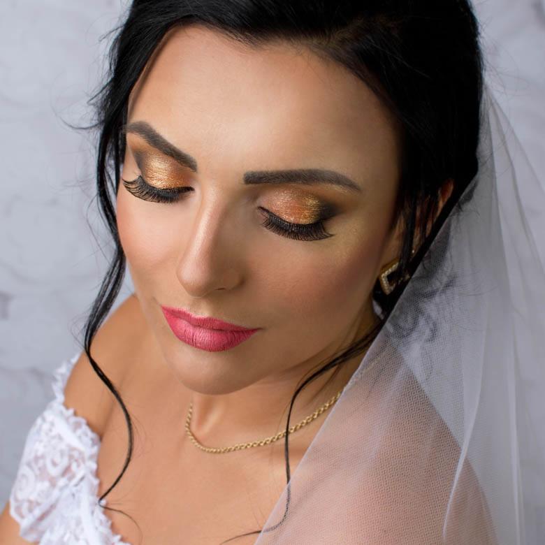 Panna młoda z zamkniętymi oczami prezentuje złoty makijaż oczu z długimi rzęsami, różowe usta i delikatną biżuterię w dniu ślubu.