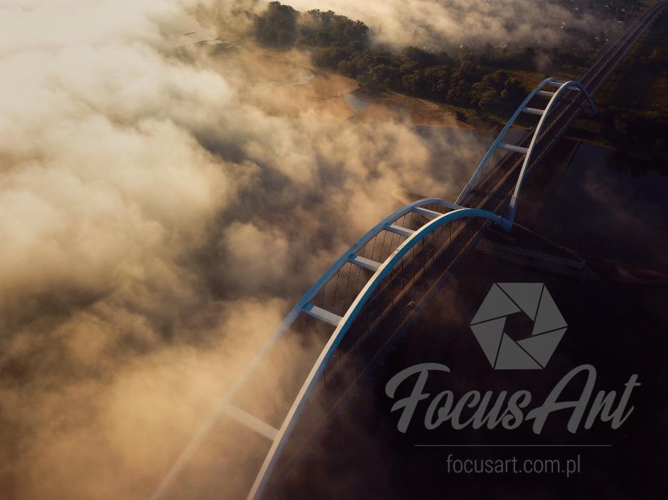 Panoramiczny widok z lotu ptaka na most łukowy zanurzony we mgle, z logo firmy Focus Art w prawym dolnym rogu.