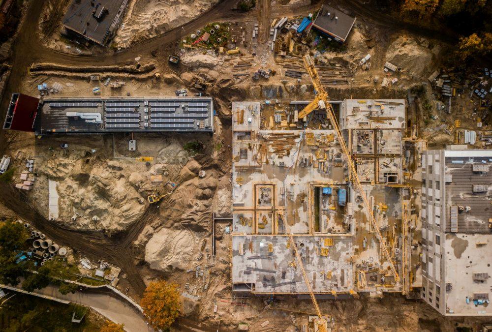 Widok z lotu ptaka na plac budowy z fundamentami budynku, żurawiami i budynkiem z panelami słonecznymi na dachu w tle, otoczony ziemią i drzewami.