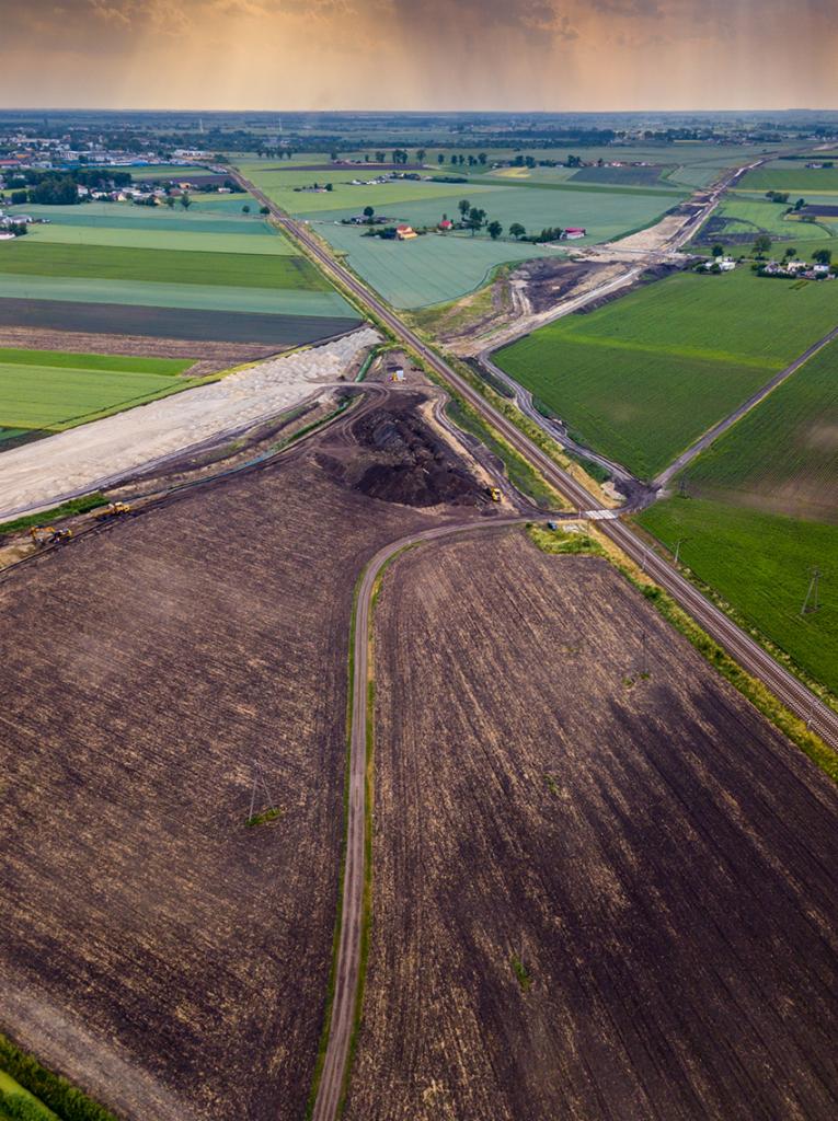 Zdjęcie z drona przedstawiające krajobraz rolniczy z budową infrastruktury liniowej, widoczne pola uprawne, fragmenty lasów, oraz nowo powstającą drogę lub linię kolejową przecinającą pola,...