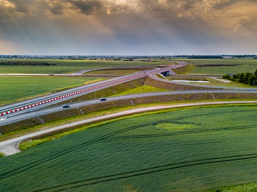 Szeroki kadr z lotu ptaka na rozległą autostradę wijącą się przez zielone pola uprawne, z dramatycznym, zachmurzonym niebem w tle.