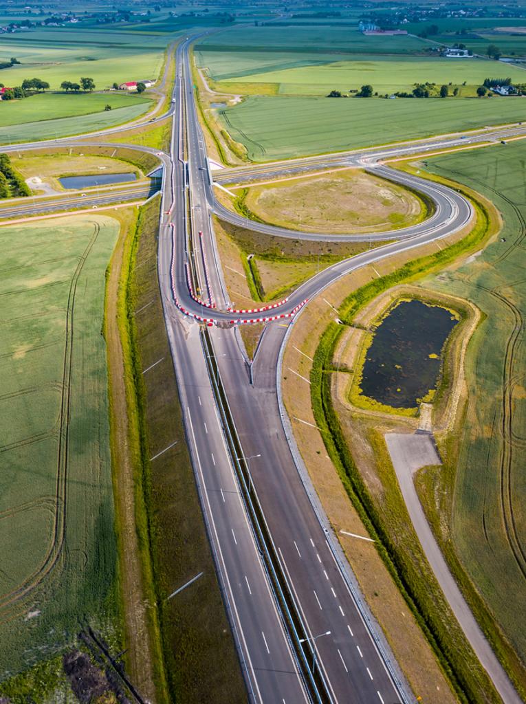 Widok z lotu ptaka na nowo wybudowaną autostradę z węzłami, otoczoną polami uprawnymi i zbiornikami retencyjnymi. Barierki ochronne w kolorze biało-czerwonym przy zjeździe. Minimalny ruch pojazdów.