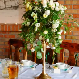 Elegancko udekorowany st&oacute;ł z wysokim, bogatym bukietem białych i kremowych r&oacute;ż oraz zieleni, zawieszonymi szklanymi kulami ze świecami, nakryty białą zastawą i lnianym obrusem, w tle ceglana ściana...